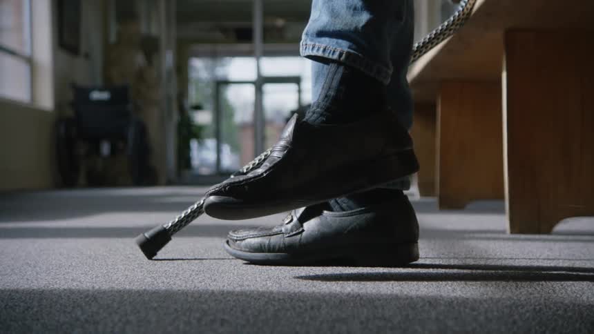 A person waiting while sitting on a bench with a cane next to them gently tapping their foot on the ground as a young girl runs back and forth in front of them