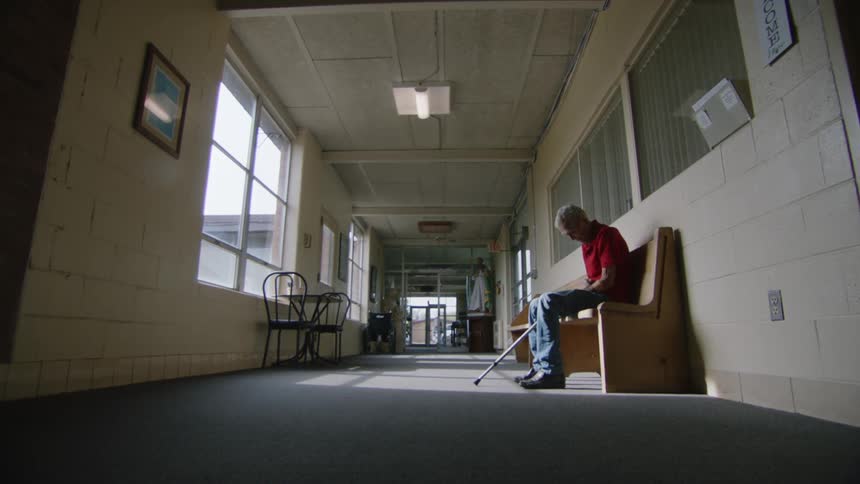 A wide shot of an elderly man with a walking stick sitting alone on a wooden bench in a long sunlit hallway