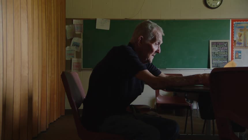 An elderly man with white hair and pale skin sits in a dark room with a green blackboard behind him leaning forward with his hands resting on a table as he looks down and to the right