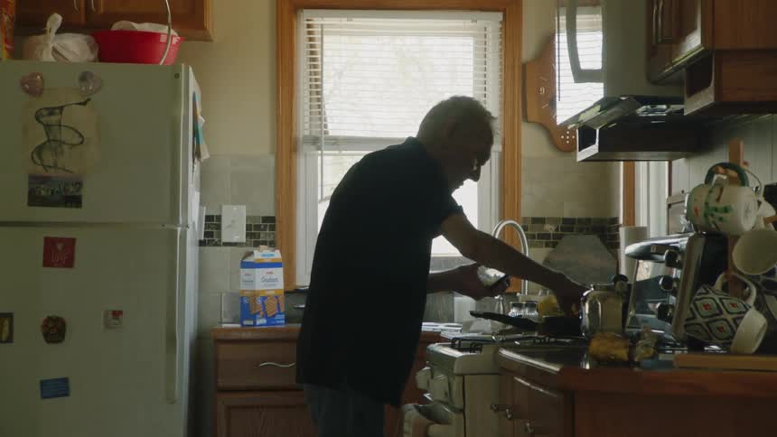 An old man is standing in his kitchen at the stove dumping food into a pan to cook