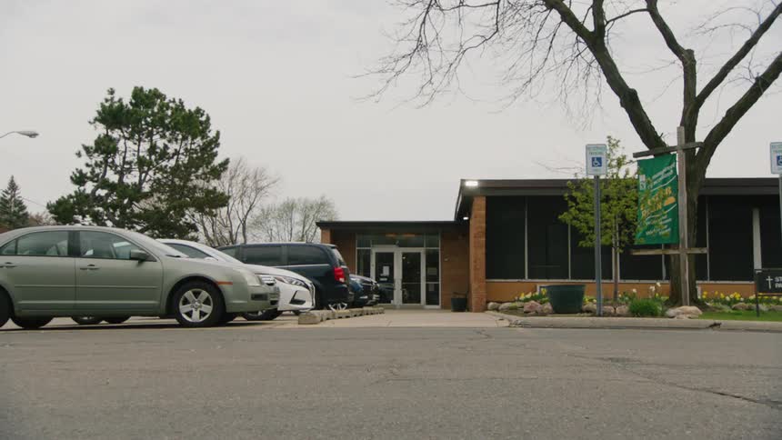 A static eyelevel shot shows the brick entrance of a modern singlestory church with several cars parked in the foreground under a completely overcast sky