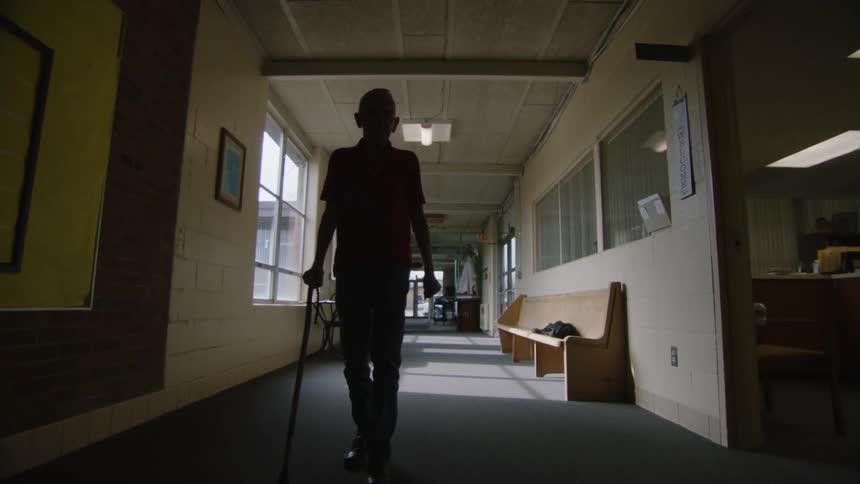 Elderly man waiting on a bench in the corridor gets up and walks with the help of a cane