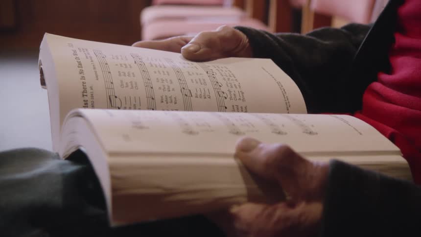 An elderly man holds a hymn book while seated inside a church