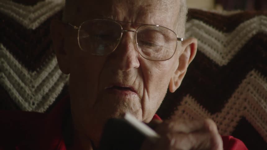 A closeup shot of an elderly man with glasses showing him holding a lit cigarette and a smartphone