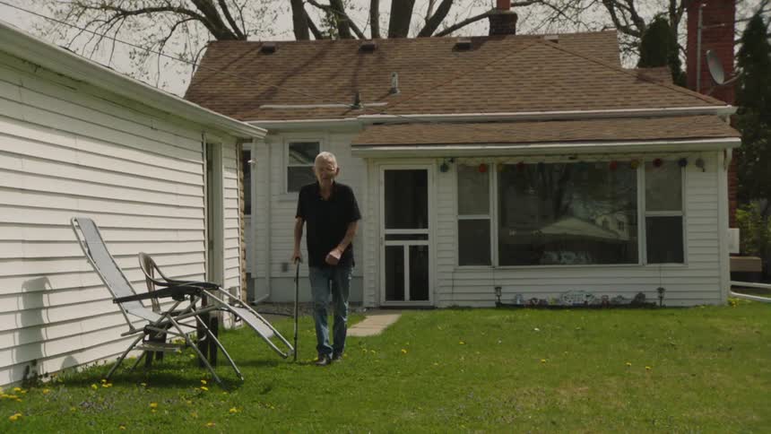 The video shows a man with white hair and a white beard walking in his backyard he is using a cane and appears to have a slight limp as he moves toward a couple of folding chairs