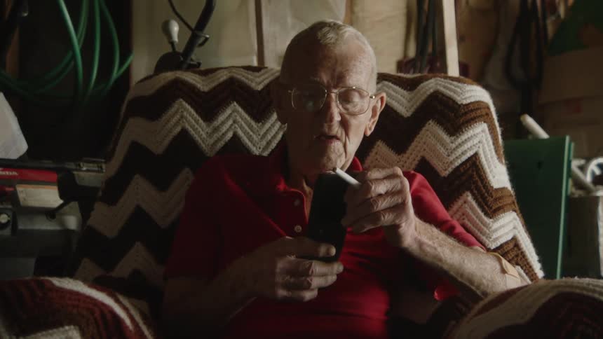Elderly man sits in a cluttered garage holding a cigarette