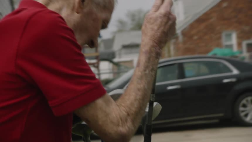 The video shows a man with white hair wearing a red polo shirt walking outside in what appears to be a residential neighborhood he is looking down at the ground as he walks with houses and cars visible in the background