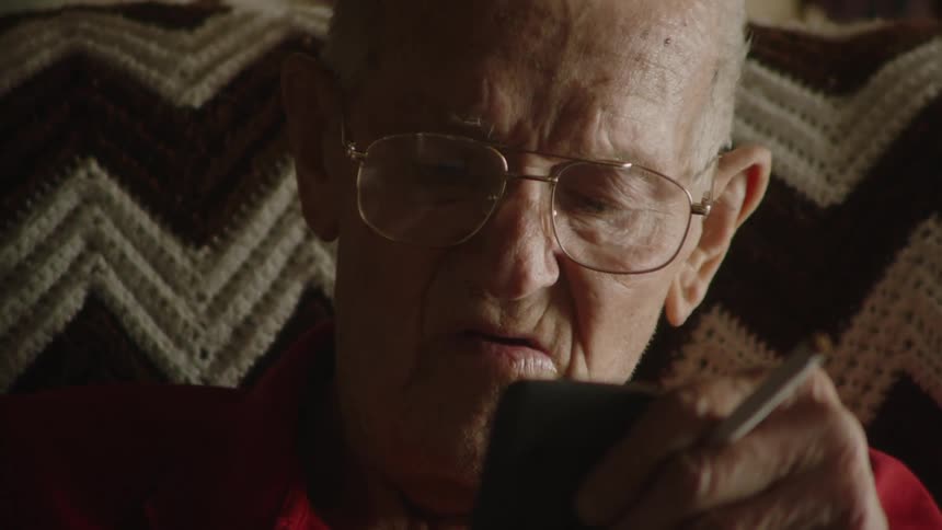 A closeup of an elderly man with glasses looking down at his phone he is holding a cigarette between his fingers