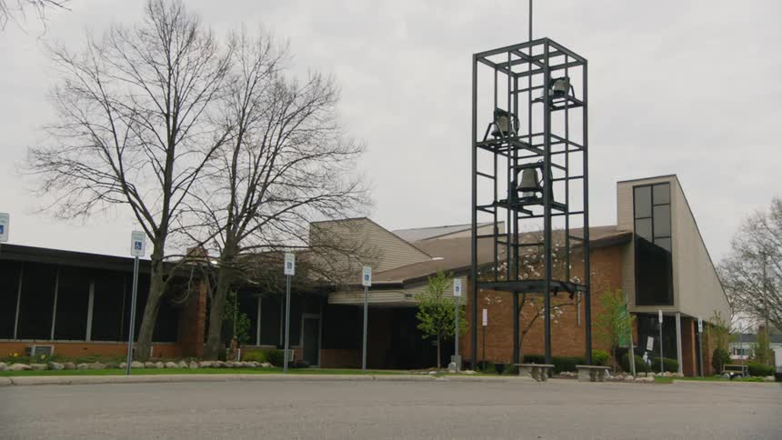 A wide shot of a modernlooking brick church building on a cloudy day in front of the church there is a large black metal structure holding three bells