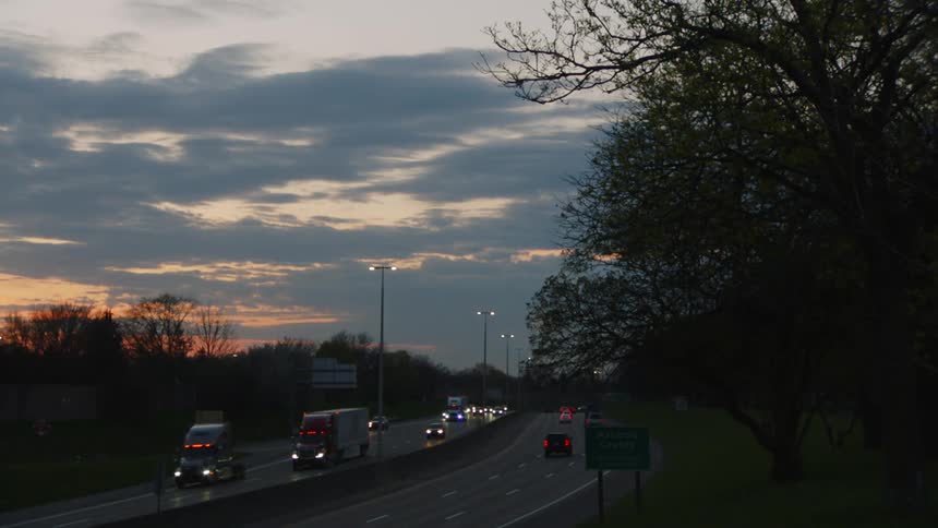 A view of a busy highway at dusk with cars and trucks traveling in both directions the sky is filled with dark clouds but a vibrant orange and pink sunset is visible on the horizon
