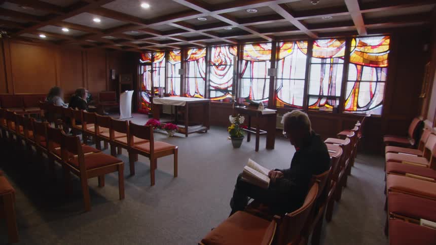 An elderly man sits on a chair in a church reading a bible with two other people seated farther away in another row