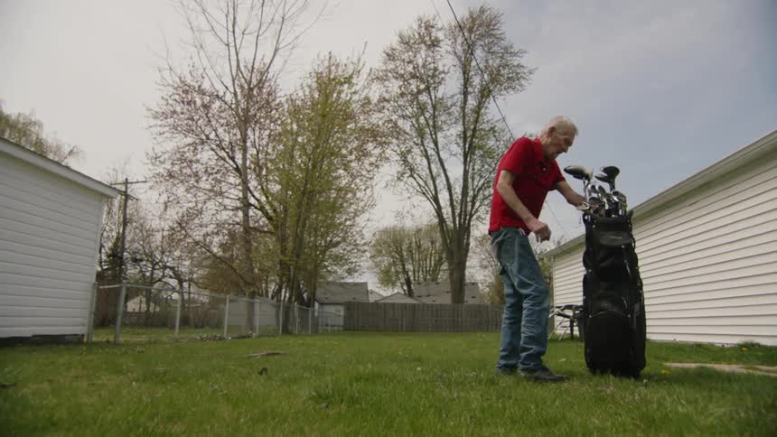 An elderly man picks up a golf club in his yard and puts in it the golf bag