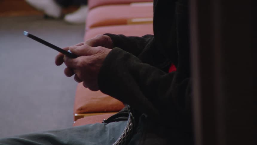 An elderly man wearing glasses scrolls through his phone while seated at one end of a row of chairs with a young man sitting at the other end