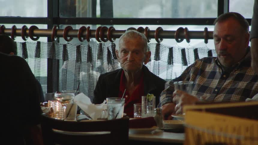 A group of adult men have drinks at a restaurant at midday while other customers walk by