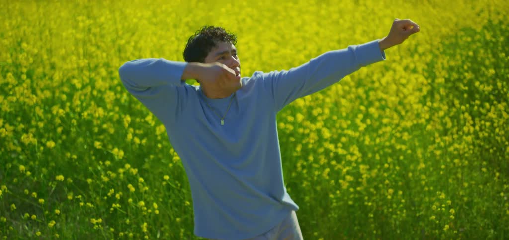 In the video a man with curly brown hair wearing a light blue longsleeved shirt and gray pants is performing a sequence of slow deliberate dance movements in the middle of a field of vibrant yellow flowers the camera focuses on him from the waist up