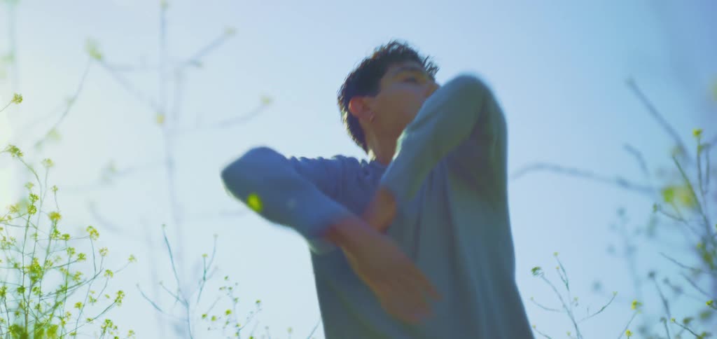 Against a bright blue sky a man in a light blue shirt performs a slow expressive dance the camera is positioned at a low angle capturing his deliberate arm movements as sunlight creates a soft halo effect around him