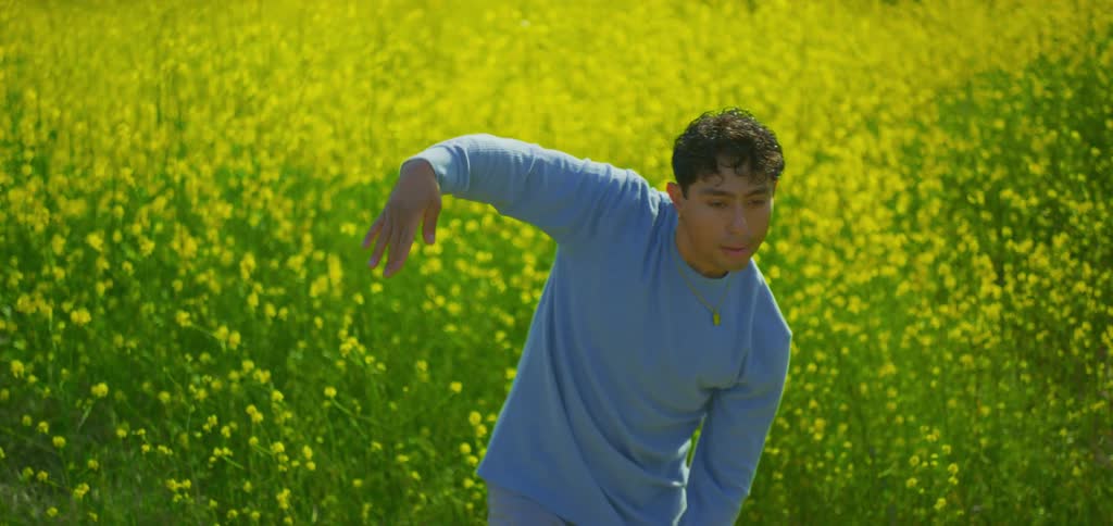 A man in a light blue longsleeved shirt is dancing in a wide sunlit field of yellow flowers he performs slow and intentional hand movements his upper body swaying gently to the rhythm the bright yellow of the flowers fills the frame creating a vibra