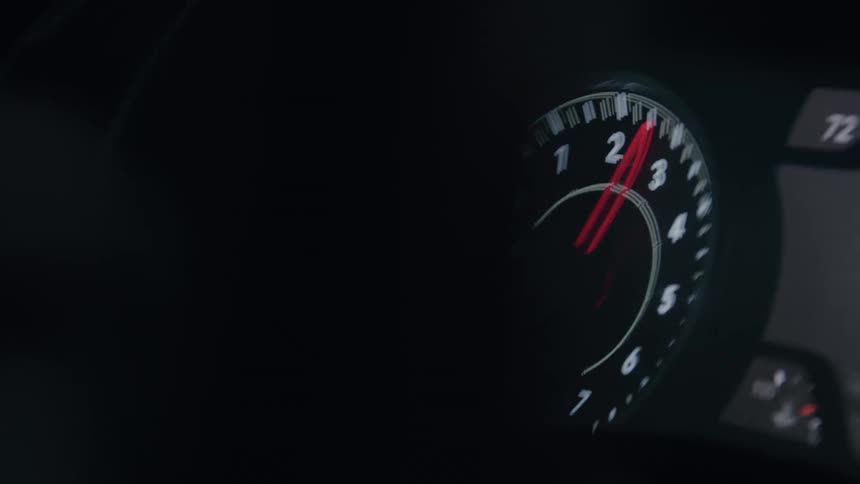 A black automobile tachometer glows with white numbers and markings as a red needle rapidly swings in between