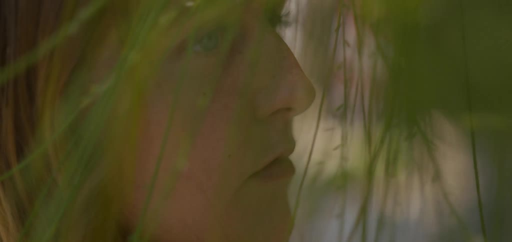 The video shows a closeup of a womans profile with a soft focus on her face and light blue eyes as she looks offscreen the foreground is partially obscured by green leaves and thin branches creating a gentle natural frame around her