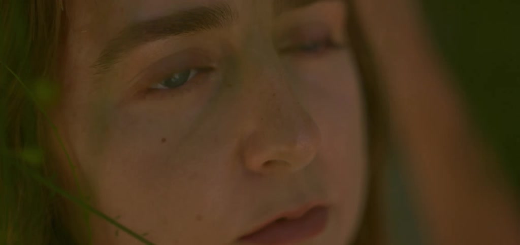 In an extreme closeup the face of a sad woman with blue eyes is partially obscured by grass as she looks up and a single tear rolls down her cheek
