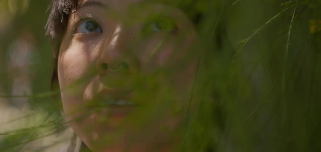 The video shows a closeup shot of a woman looking up and smiling with her face partially obscured by green leaves and branches in the foreground the camera focuses on her peaceful expression as she looks upwards framed by the surrounding foliage