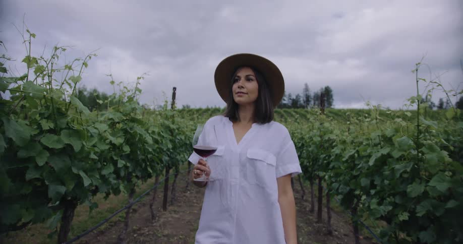 A woman holding a wine glass walks through rows of grapevines in a vineyard