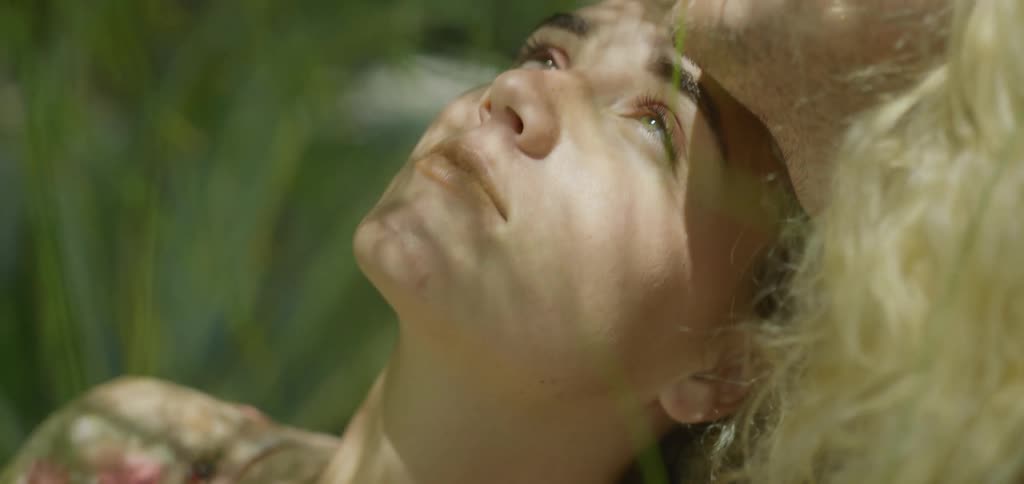 The video shows an intimate closeup of a couple looking up at the sky the woman with her head resting on the mans chest looks peaceful while he has a thoughtful expression as they both enjoy the dappled sunlight filtering through the surrounding pla