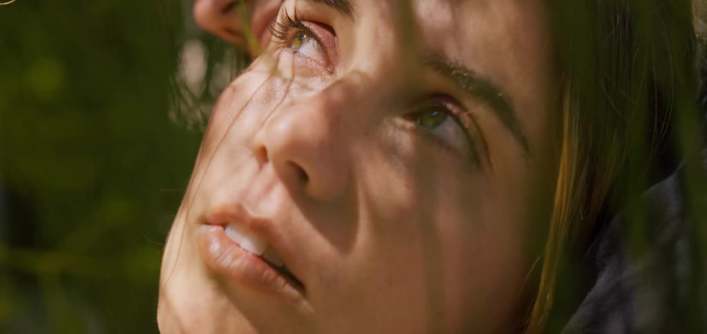 The video shows an extreme closeup of a womans face she has her eyes closed and a peaceful serene expression while dappled sunlight and the shadows of leaves flicker across her features