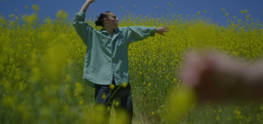 A young woman wearing an oversized shirt and trousers dances in an open tall grass field