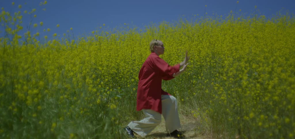 A young woman wearing an oversized red shirt and milk trousers dances in an open tall grass field