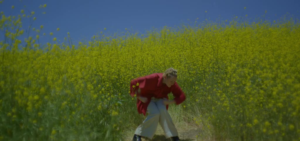 A young woman wearing an oversized shirt and trousers dances in an open tall grass field