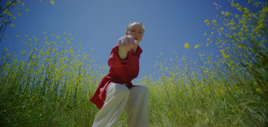 Blonde woman dancing outdoors among a field of yellow flowers against a blue sky
