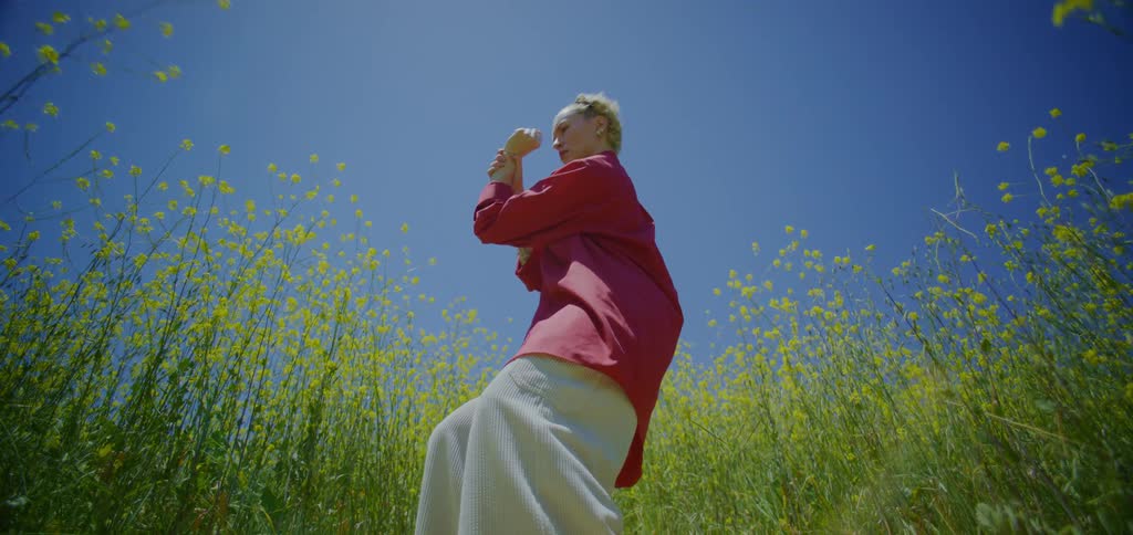 A lowangle shot of a woman in a red shirt and white pants joyfully dancing with her arms outstretched in a field of yellow flowers under a clear blue sky