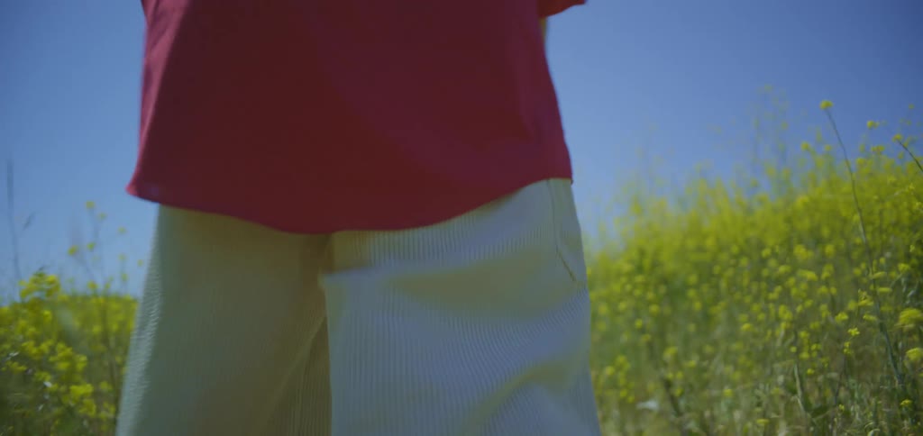 A shot from the knees down shows a person walking through a field of yellow wildflowers the person is wearing a long red shirt and lightcolored pants
