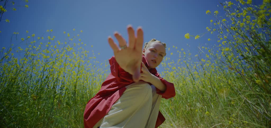 The video shows a lowangle shot of a person in a red shirt and white pants performing a slow expressive dance they are crouching down on a dirt path surrounded by a field of tall yellow flowers under a clear blue sky