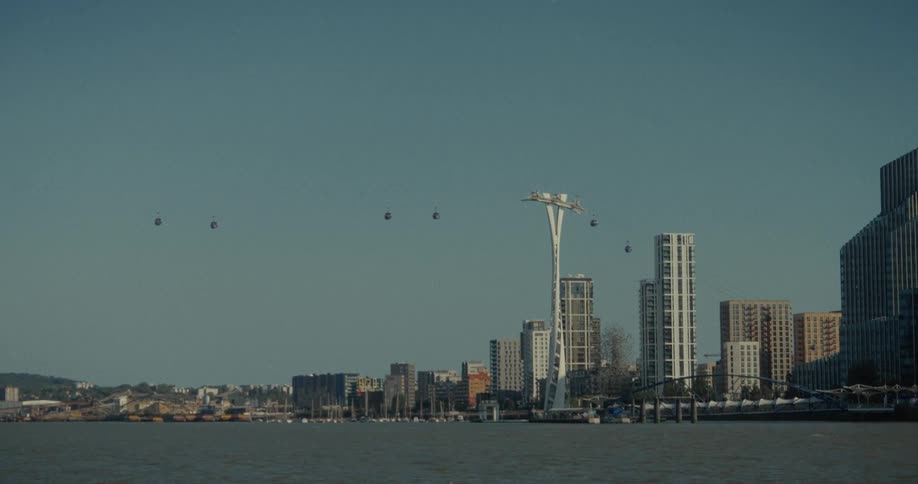 A distant view of a city on a waterfront shows a large body of water in the foreground and a skyline of tall modern buildings and docked boats against a clear blue sky with several cable cars suspended in the air