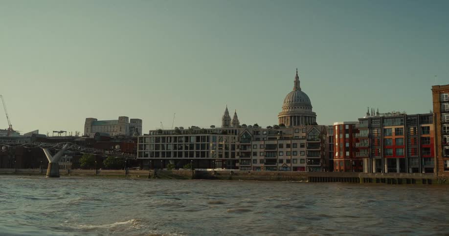 A long view from across a wide river shows a white stone cathedral with a large dome rising above a row of brick buildings on the right and a modern steel bridge on the left under a pale blue sky