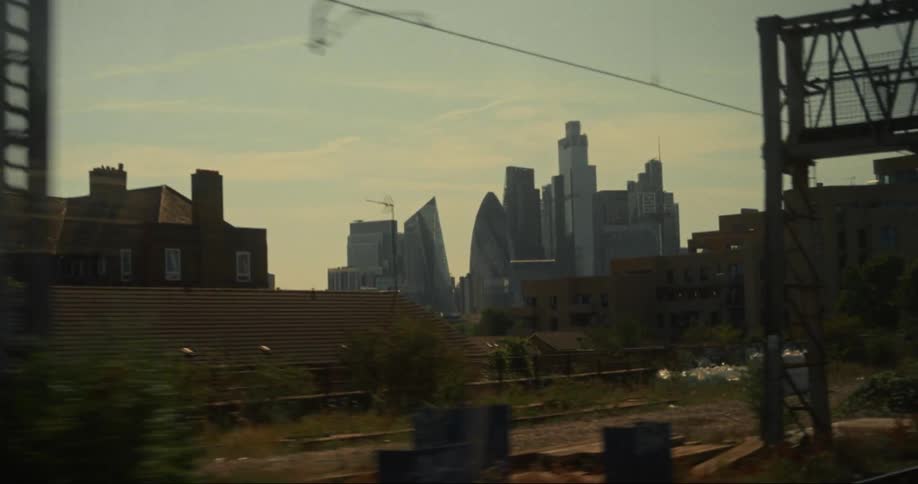 The video shows a view from a train window looking out over a city skyline the foreground features rooftops and power lines while the background is dominated by tall modern skyscrapers under a hazy sky
