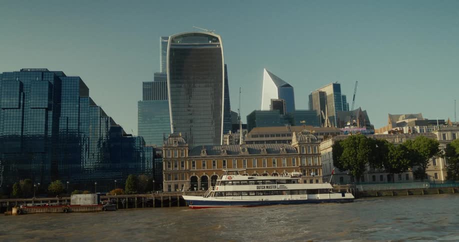 A long two story ferry with a white and blue hull and a red stripe travels across a wide muddy river with tall glass skyscrapers and a large ornate building on the far bank under a hazy blue sky