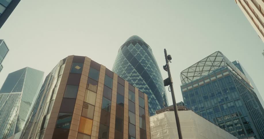 The video shows a busy city street scene the camera pans upwards from a streetlevel view of a sidewalk and a bench to reveal several tall modern buildings and a glass skyscraper