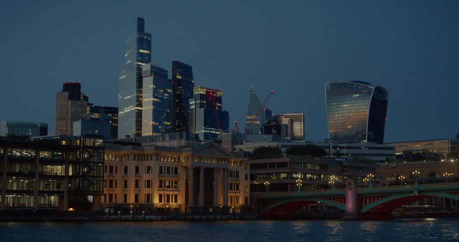 A view of a large city skyline at night with multiple tall modern buildings and a bright red bridge with lights illuminating the windows of the buildings and reflected on the water below