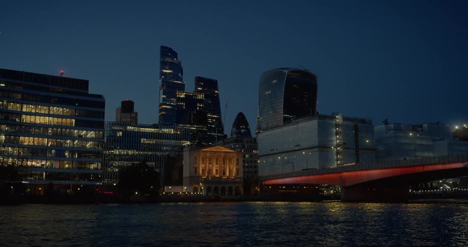 A large city skyline with several tall modern buildings and a bright red bridge is visible from a body of water at night