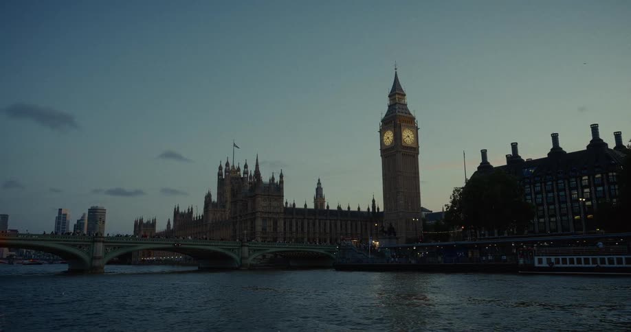 A wide view of london at dusk shows the houses of parliament and the big ben clock tower with its faces lit up all standing beside a wide river with a busy bridge and city skyline visible on the left