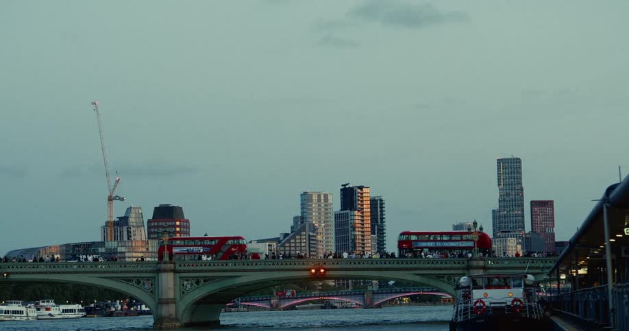 A bridge connecting two parts of a city with doubledecker buses passing by and a large crowd of people lining the sidewalks and a series of highrise buildings in the background at midday