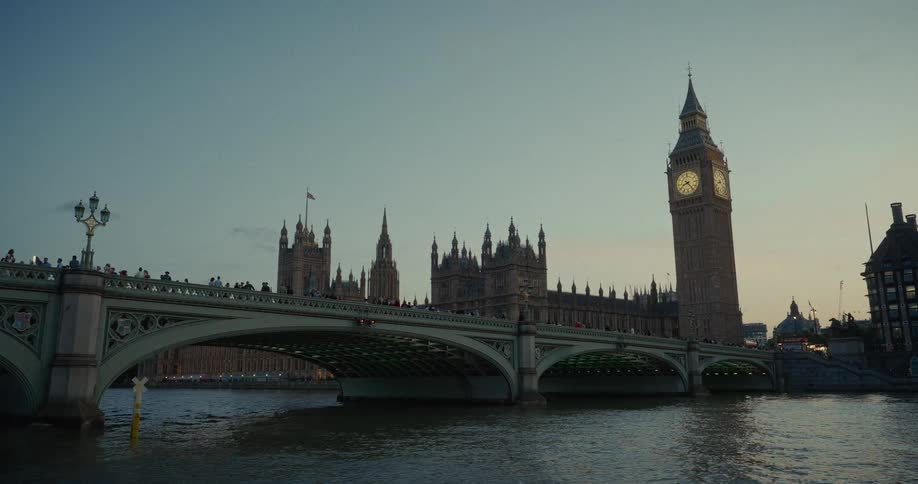 A view across the water shows the houses of parliament and big ben clock tower with its lit up face as people walk along the top of a wide bridge in the foreground
