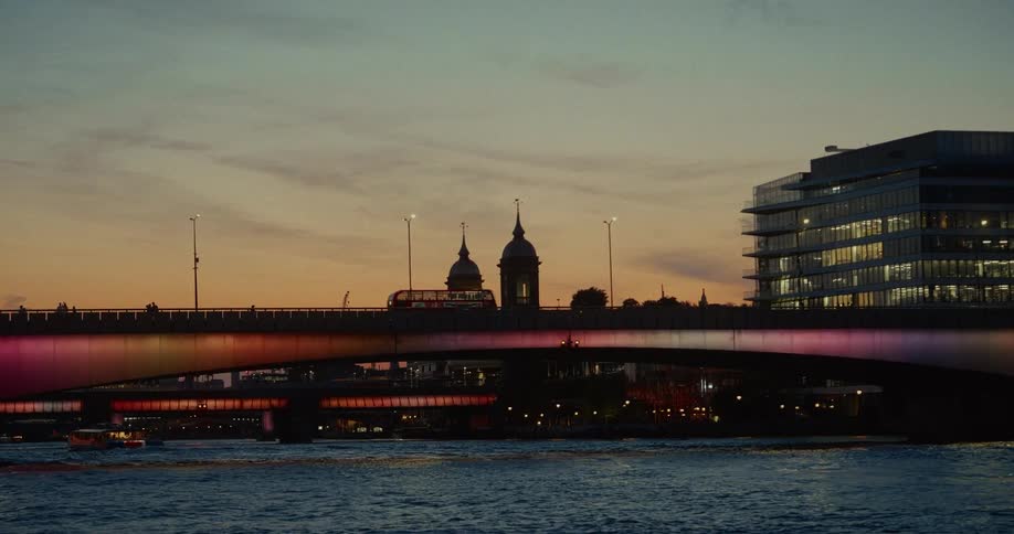 A wide shot captures a city skyline at dusk with a bridge illuminated by colorful lights a doubledecker bus crossing it and domed buildings in the background