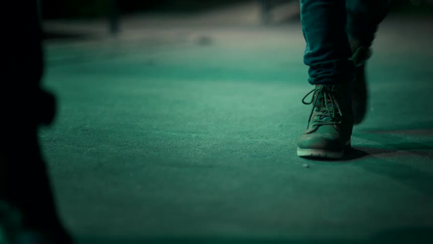 Two people wearing dark boots face each other on a street lit by green light