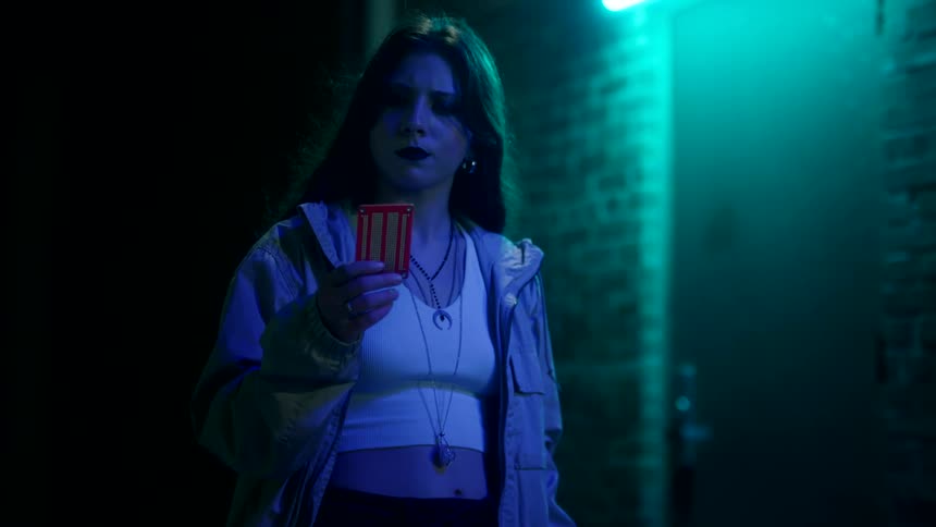 Young woman looks at a red card in an alley lit by blue neon light