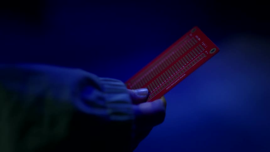 The video shows a hand holding a red circuit board against a dark blue background the scene is lit with a blue light that creates a shadowy almost mysterious atmosphere