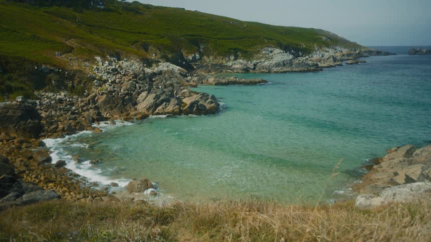 Surf flows onto the rocky shore at the base of a grassy slope on a summer day